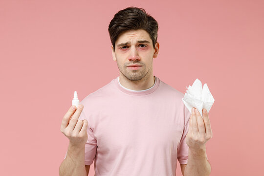 Frustrated sick unhealthy ill allergic man has red eyes runny stuffy sore nose suffer from allergy symptoms hay fever hold paper napkin use nasal drops isolated on pastel pink color background studio