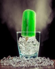A green mint popsicle is standing inside a glass with ice and smoke on the dark background