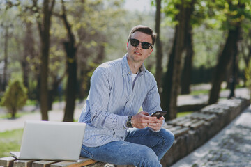 Side view young thoughtful pensive caucasian freelancer man in shirt glasses sit on park bench work by laptop pc computer outdoors, using mobile cell phone wi-fi look aside Concept of urban lifestyle.
