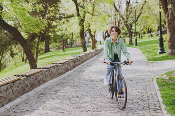 Young fun smiling happy woman 20s wearing casual green jacket jeans riding bicycle bike on sidewalk...