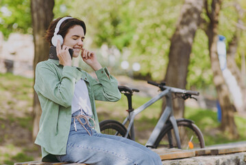 Fototapeta na wymiar Young happy fun woman in casual green jacket jeans headphones sit on bench near bicycle bike in city spring park outdoors hold mobile cell phone listen to music. People active urban youthful concept.
