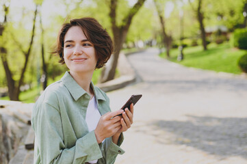Young pensive dreamful smiling woman in casual green jacket jeans sit on bench in city spring park outdoors resting use mobile cell phone chat online look aside. People active urban lifestyle concept.