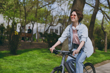 Side view young satisfied happy sporty woman in jeans clothes headphones riding bicycle bike on...