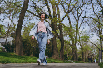 Full length bottom view young smiling fun confident woman 20s wearing trendy jeans clothes walking strolling in spring green park outdoors go down alley, looking camera. People urban lifestyle concept