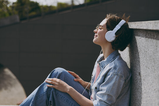 Young Side View Smiling Caucasian Woman In Jeans Clothes Headphones Eyeglasses Listen To Music Leaning On Building Wall Sit On Concrete Steps Outdoors Walk Look Aside People Urban Lifestyle Concept.