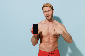Fototapeta premium Handsome young sexy man in red shorts swimsuit relax near hotel pool point finger on mobile cell phone blank screen workspace area isolated on blue background Summer vacation sea rest sun tan concept