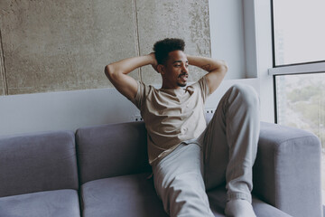 Dreamful young african american man in beige t-shirt sweatpants sit on comfortbale grey sofa indoors apartment look aside at window hold hands behind neck procrastinate, rest on weekends stay at home