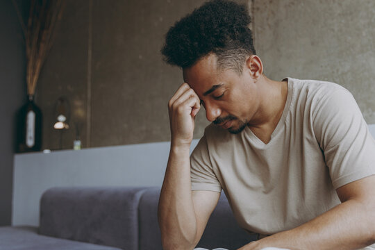 Side view sad puzzled thoughtful young african american man wear beige t-shirt sit on grey sofa indoors apartment procrastinate prop up forehead rest on weekends stay at home. Tattoo translate fun