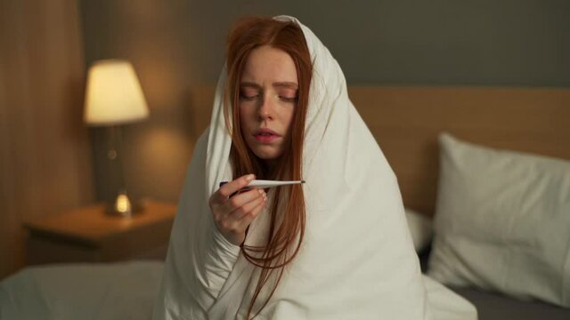 Sad lll young woman suffering from runny nose disease sitting in bed covered with warm blanket. Sick unhappy redhead female suffering from stuffy nose problem measuring temperature with thermometer