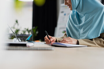 partial view of muslim businesswoman writing in notebook on blurred foreground