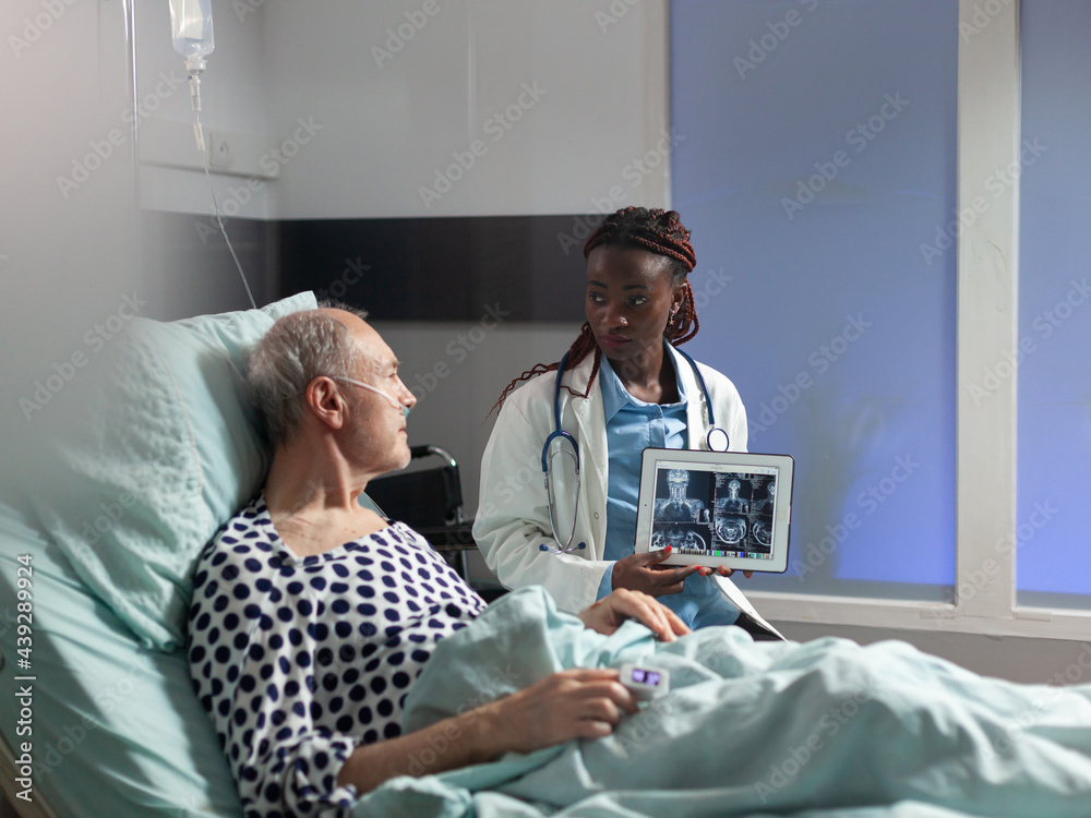 Wall mural African american doctor sitting next to senior man explaining body trauma diagnosis, showing x-ray on tablet pc in hospital room. Mature patient in clinic breathing it help from oxygen mask.