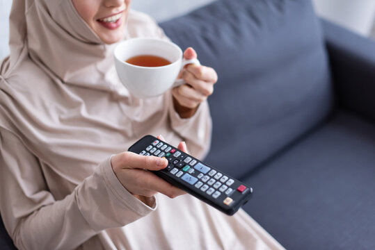 cropped view of smiling muslim woman with cup of tea and remote controller watching tv at home