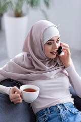 young arabian woman in hijab talking on smartphone while holding cup of tea