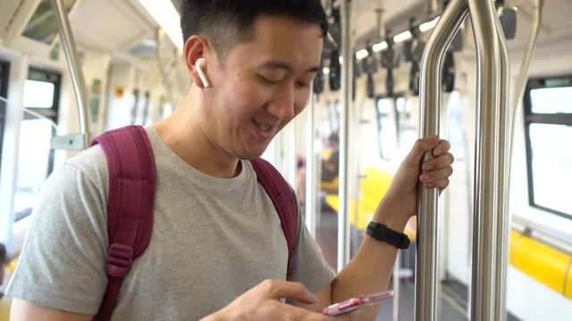 Close up of young man listening to music with wireless earpods and using mobile phone while commuting by train. Asian guy enjoying music on the go.