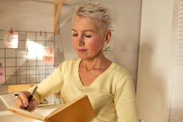 Concentrated mature blonde female working from home sitting at table handwriting in notebook, putting down her ideas and thoughts, setting goals, making plans, having focused facial expression