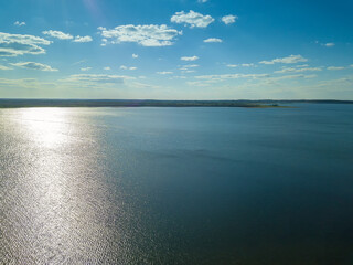 View from the drone is of a blue lake with a reflection of the sun and a green forest in the distance. Beautiful landscape from a drone on a sunny day
