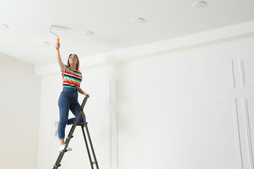 Young woman painting ceiling with white dye indoors, space for text