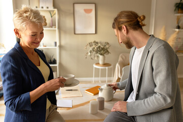 Cheerful 50 year old woman and her fashionable young male colleague sitting on desk in cozy office, drinking coffee or tea during break, speaking, sharing news, talking, having nice conversation