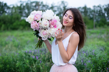 Beautiful girl with pink peonies, enjoying a bouquet of flowers on background of nature. Happy smiling woman in dress holds peonies in hands. Girl with a bouquet of flowers. Girl in wildflowers.	