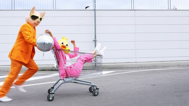 Pig man pushing bird woman with disco ball in shopping cart