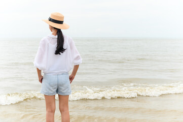 Outdoor summer portrait of young asian woman in straw hat looking to the sea at tropical beach, enjoy freedom time, fresh air and leisure travel vacation.