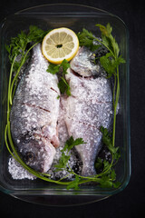 Fish in a glass tray with coarse salt, lemon slices and parsley on a dark background.