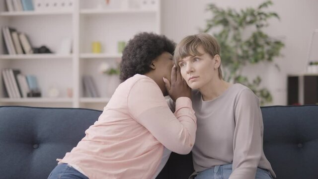 Confused african woman whispering secret to caucasian female friend, trust