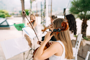 Girl musician from the string orchestra sits near the music stand and holds a cello during the wedding ceremony, back view 