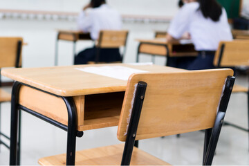 Class room tables and chairs with Paper documents of exam test on desk in examination school while blur asian girls students taking reading for testing in classroom school, Student uniform in Thailand
