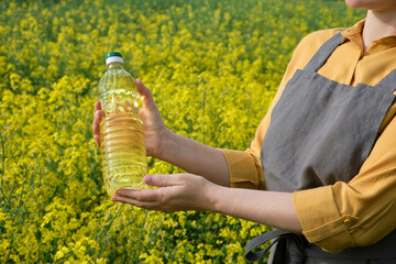 A woman stands in a rape field and holds a bottle of rapeseed oil in his hands. Concept of canola oil production. - obrazy, fototapety, plakaty