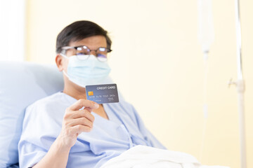 Asian male patients wear a mask and showing credit card  for medical expenses on a hospital bed. Elderly health insurance, Life Insurance and financial planning