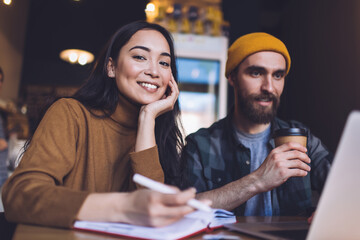 Smiling young lady taking notes sitting with coworker in cafeteria