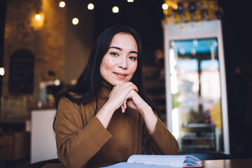 Portrait of attractive Caucasian female student looking at camera with cute smile on face, charming hipster girl sitting at cafeteria table with education notebook for learning and studying