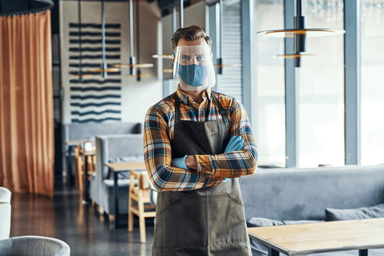 Male waiter in protective workwear keeping arms crossed and looking at camera while standing in restaurant