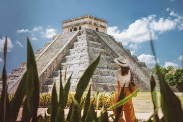 A young woman tourist in a hat stands against the background of the pyramid of Kukulcan in the ancient Mexican city of Chichen Itza. Travel concept.Mayan pyramids in Yucatan, Mexico - obrazy, fototapety, plakaty