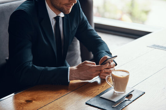 Close up of young businessman in full suit using smart phone while sitting in the restaurant