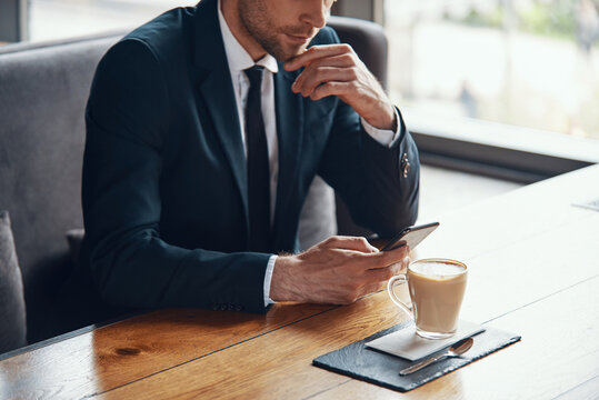 Close up of young businessman in full suit using smart phone while sitting in the restaurant