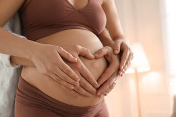 Pregnant young woman making heart with hands on belly and husband near her at home, closeup