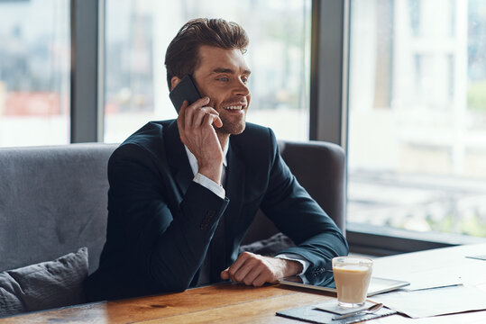 Smiling young businessman in full suit talking on the smart phone while sitting in the restaurant