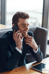 Smiling young businessman in full suit talking on the smart phone while sitting in the restaurant