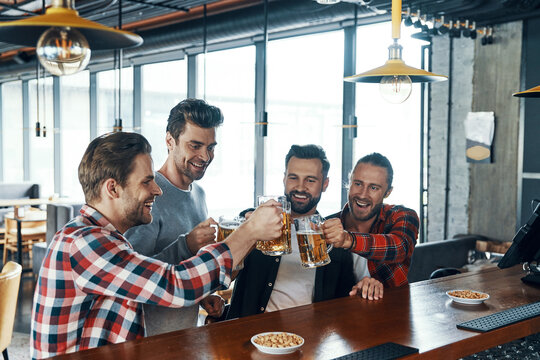 Happy young men in casual clothing toasting each other with beer and laughing while sitting in the pub