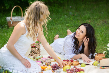Two girls are resting in park sitting on a picnic blanket. Friends is making picnic outdoor with fruits and wine. Blonde and brunette girl in nature.