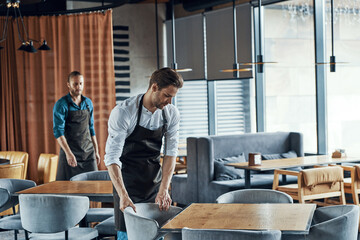 Handsome young men in aprons arranging furniture while preparing restaurant to opening