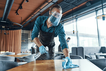 Young male waiters in protective workwear cleaning tables in restaurant