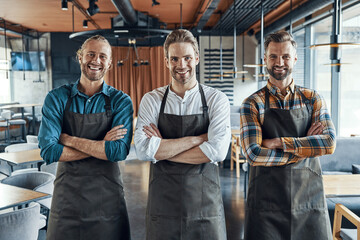 Three handsome male waiters in aprons keeping arms crossed and looking at camera