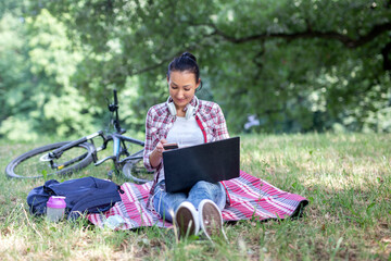 Happy young woman in casual red plaid shirt sits on red plaid picnic blanket and texts message on smartphone while working on laptop outside in park