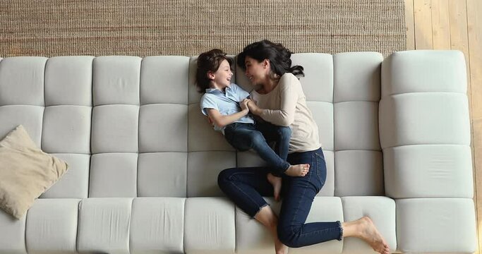 Happy Asian Mom Tickling Little Adorable Son, Above View. Cheery Lively Preschooler Boy Play On Sofa With Young Baby Sitter. Multiethnic Family Enjoy Carefree Playtime At Modern Home, Have Fun Concept