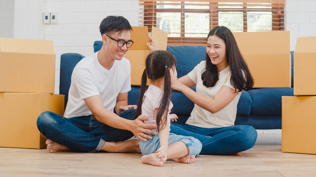 Happy Asian young family homeowners bought new house. Korean Mom, Dad, and daughter playing together during unpacking in new home after moving in relocation sitting on floor with boxes together.