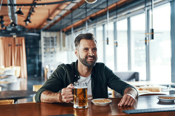 Happy young man in casual clothing drinking beer while spending time in the pub