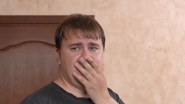 Emotional scared young plump man shakes head with hard shock closing open mouth by palm at cabinet in light room at home closeup